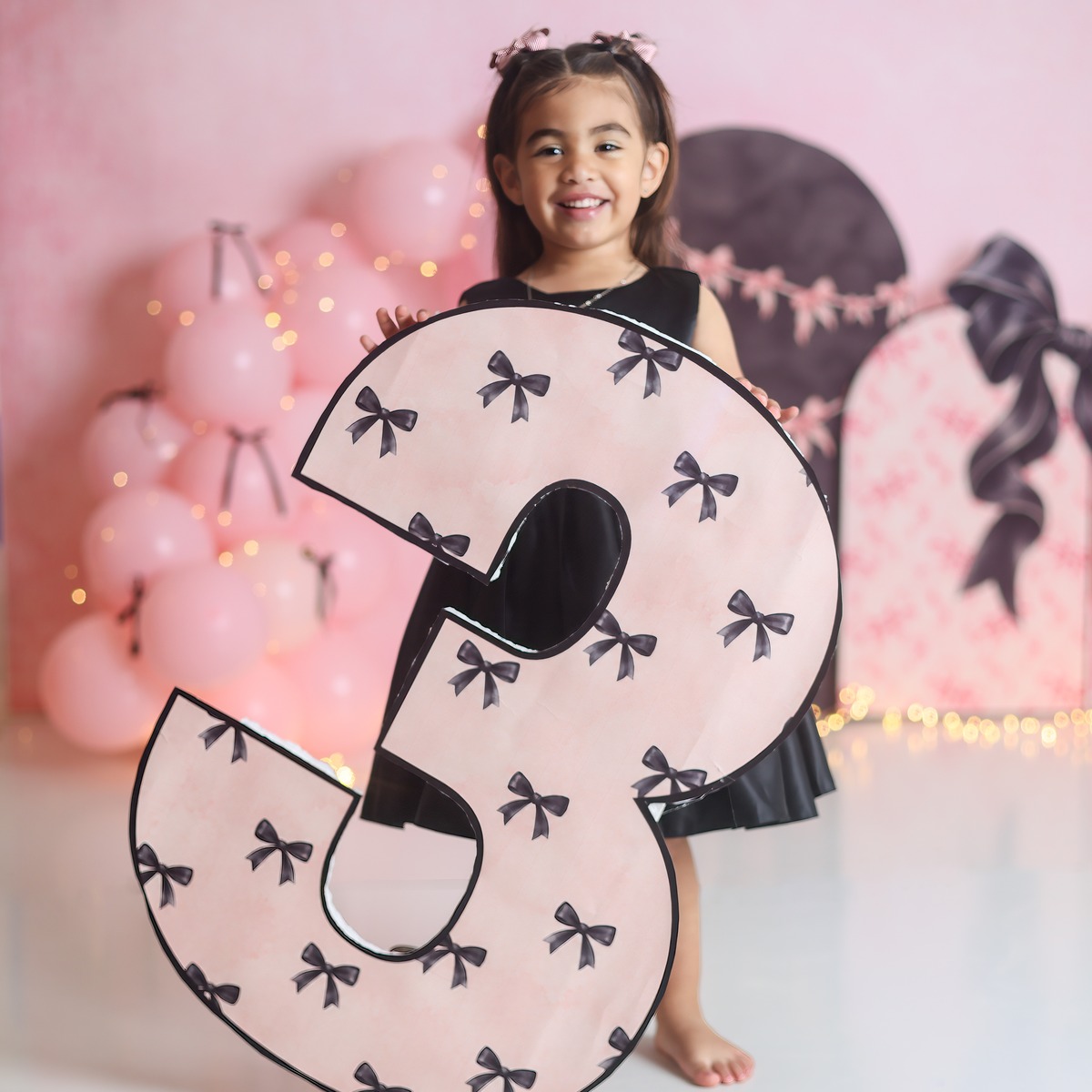 A smiling child holding a pink number three with black bows