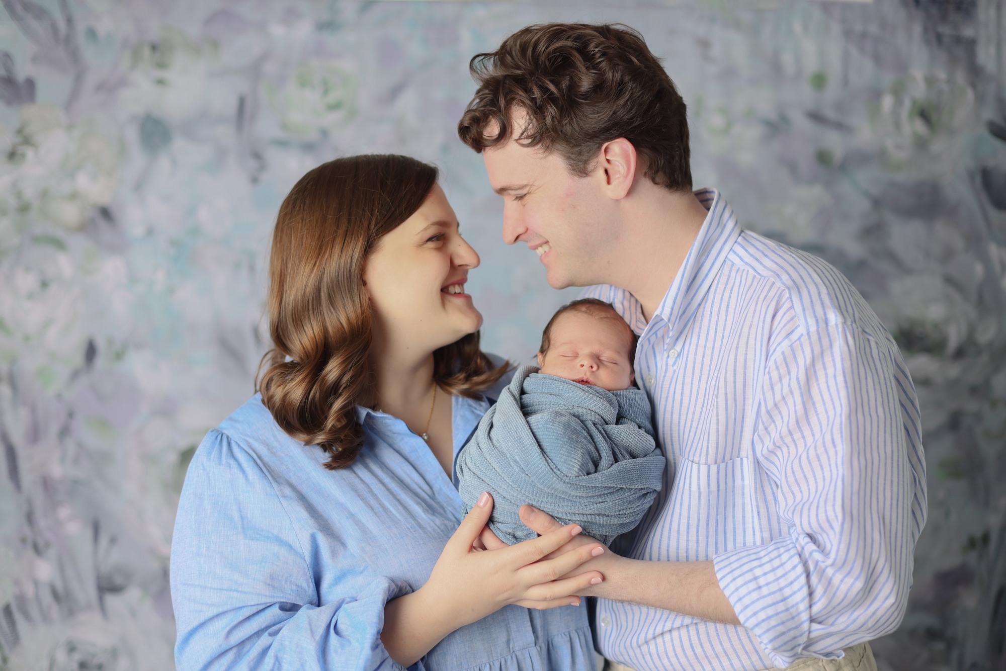 Parents smiling at each other while cradling their sleeping newborn in soft blue