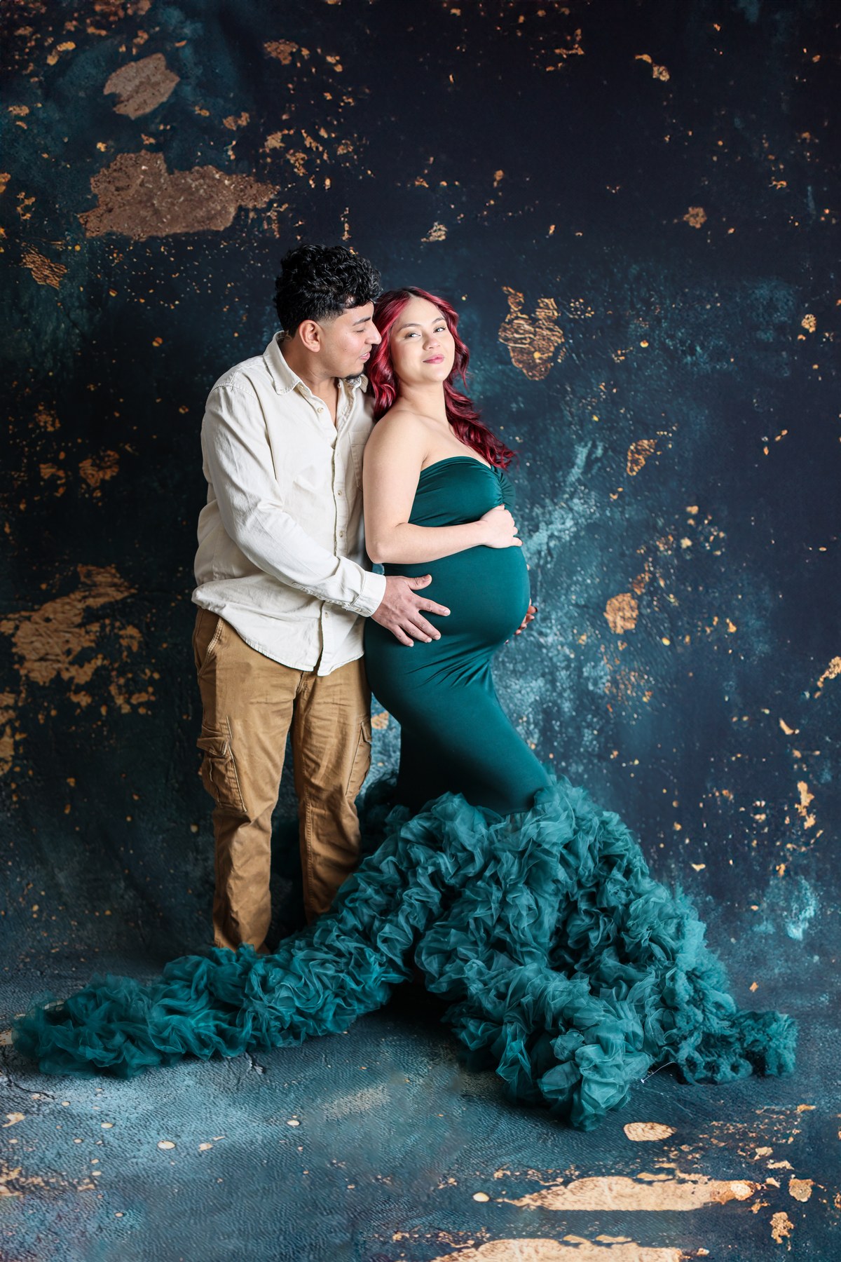 Expectant parents sharing a kiss in a teal gown against a blue and gold backdrop