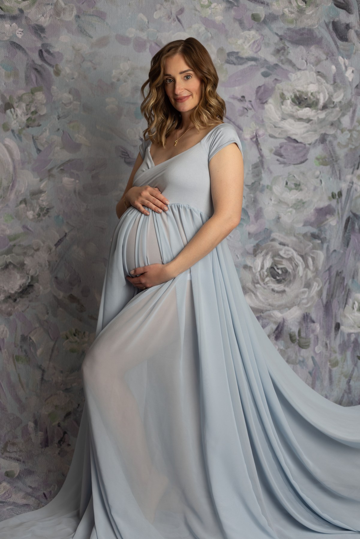 Maternity portrait in a pale blue gown against a soft floral studio backdrop