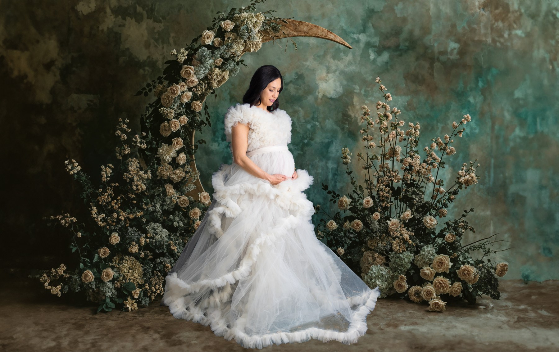 An expectant mother looking down at her bump in a white tulle gown beside a floral crescent against a painterly green backdrop