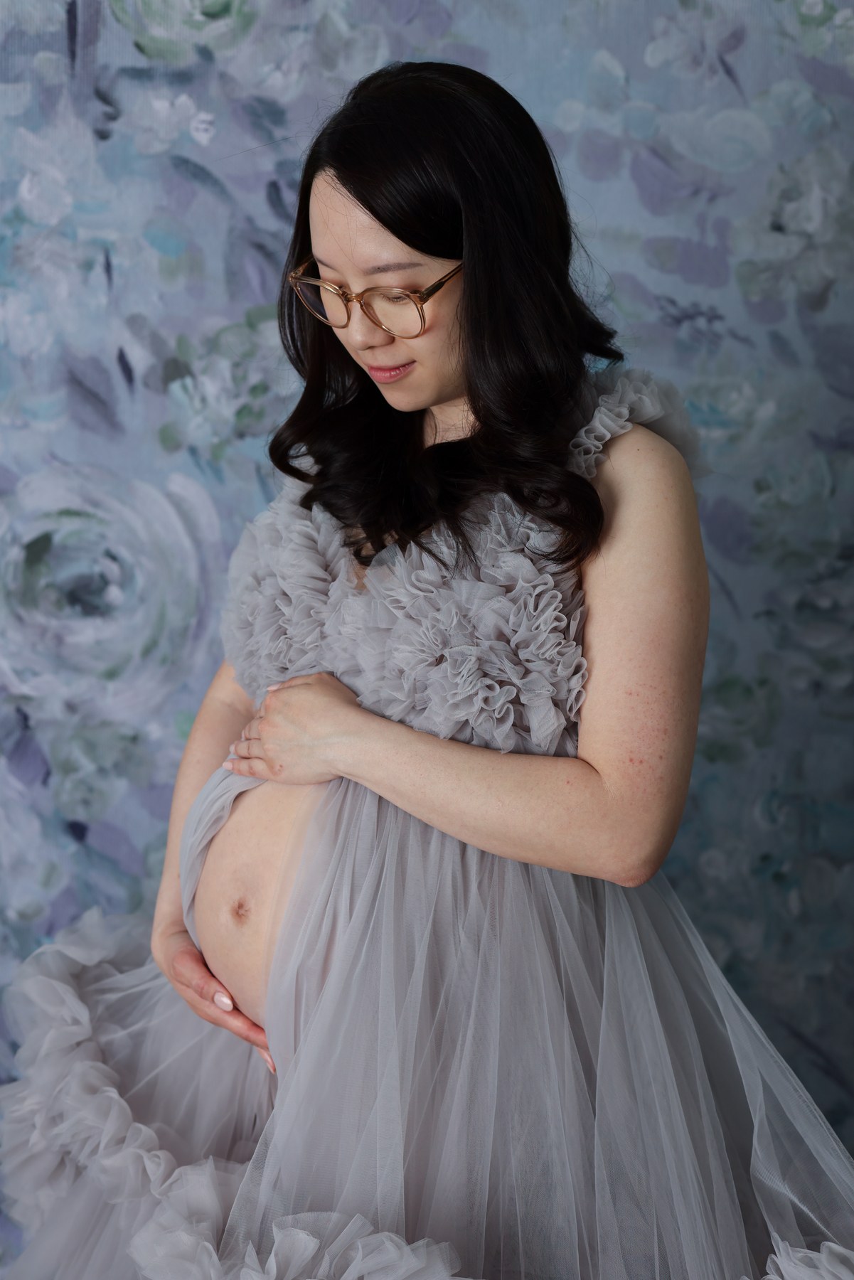 An expectant mother holding her bump in a lavender gown against blue flowers