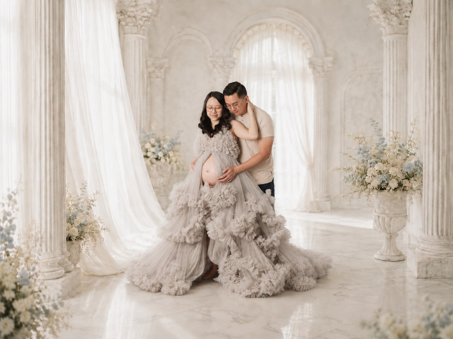 Expectant parents embracing in a pale ruffled gown in an ornate light-filled setting