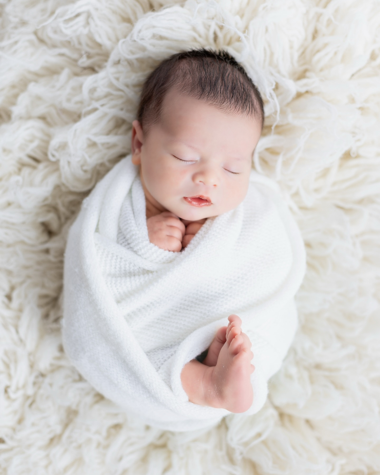 A sleeping newborn wrapped in ivory with one hand beside the cheek and tiny toes visible on a cream blanket